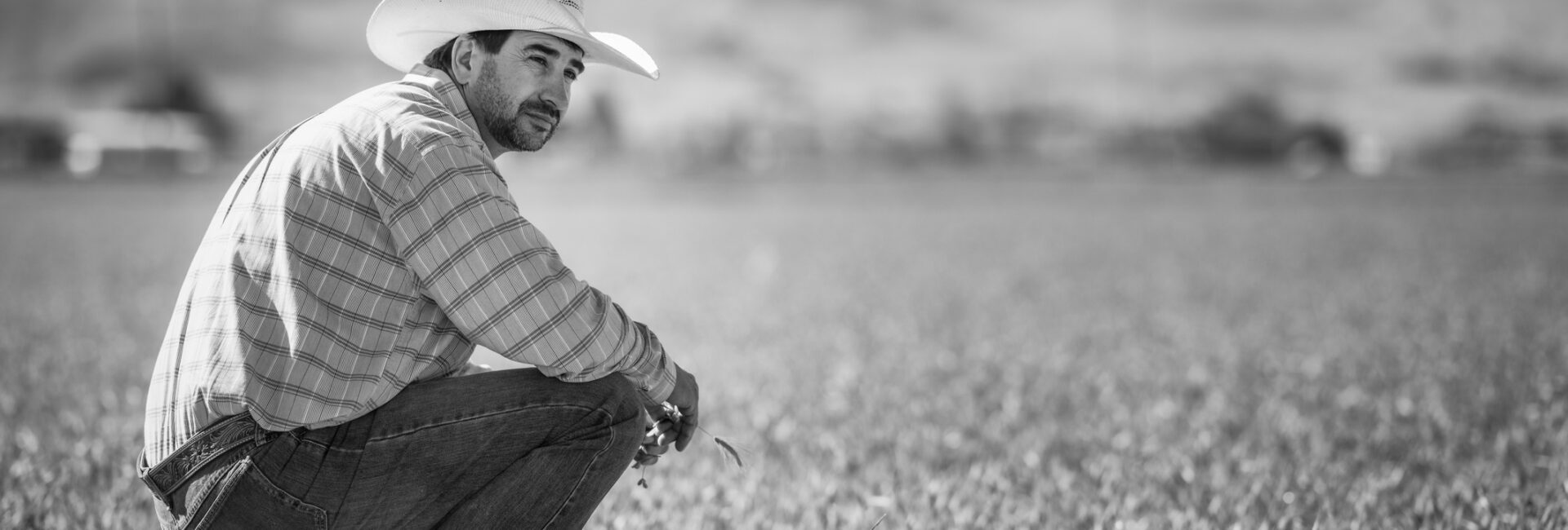 A man in a cowboy hat and plaid shirt squats in a grassy field with mountains behind him, capturing the mental load of agriculture in black and white. For support, reach out to an ag hotline.
