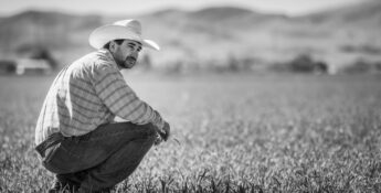 A man in a cowboy hat and plaid shirt squats in a grassy field with mountains behind him, capturing the mental load of agriculture in black and white. For support, reach out to an ag hotline.