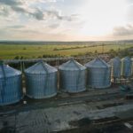 A row of large metal grain silos stands beside railway tracks, surrounded by open fields under a partly cloudy sky, as the sun sets—an ideal setting for leveraging the 45Z clean fuel production credit in sustainable agriculture.