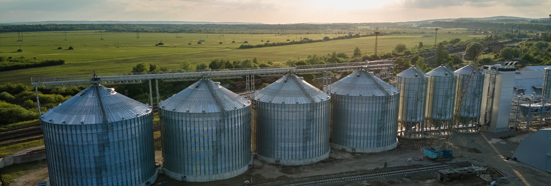 A row of large metal grain silos stands beside railway tracks, surrounded by open fields under a partly cloudy sky, as the sun sets—an ideal setting for leveraging the 45Z clean fuel production credit in sustainable agriculture.