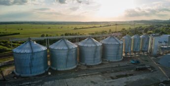 A row of large metal grain silos stands beside railway tracks, surrounded by open fields under a partly cloudy sky, as the sun sets—an ideal setting for leveraging the 45Z clean fuel production credit in sustainable agriculture.