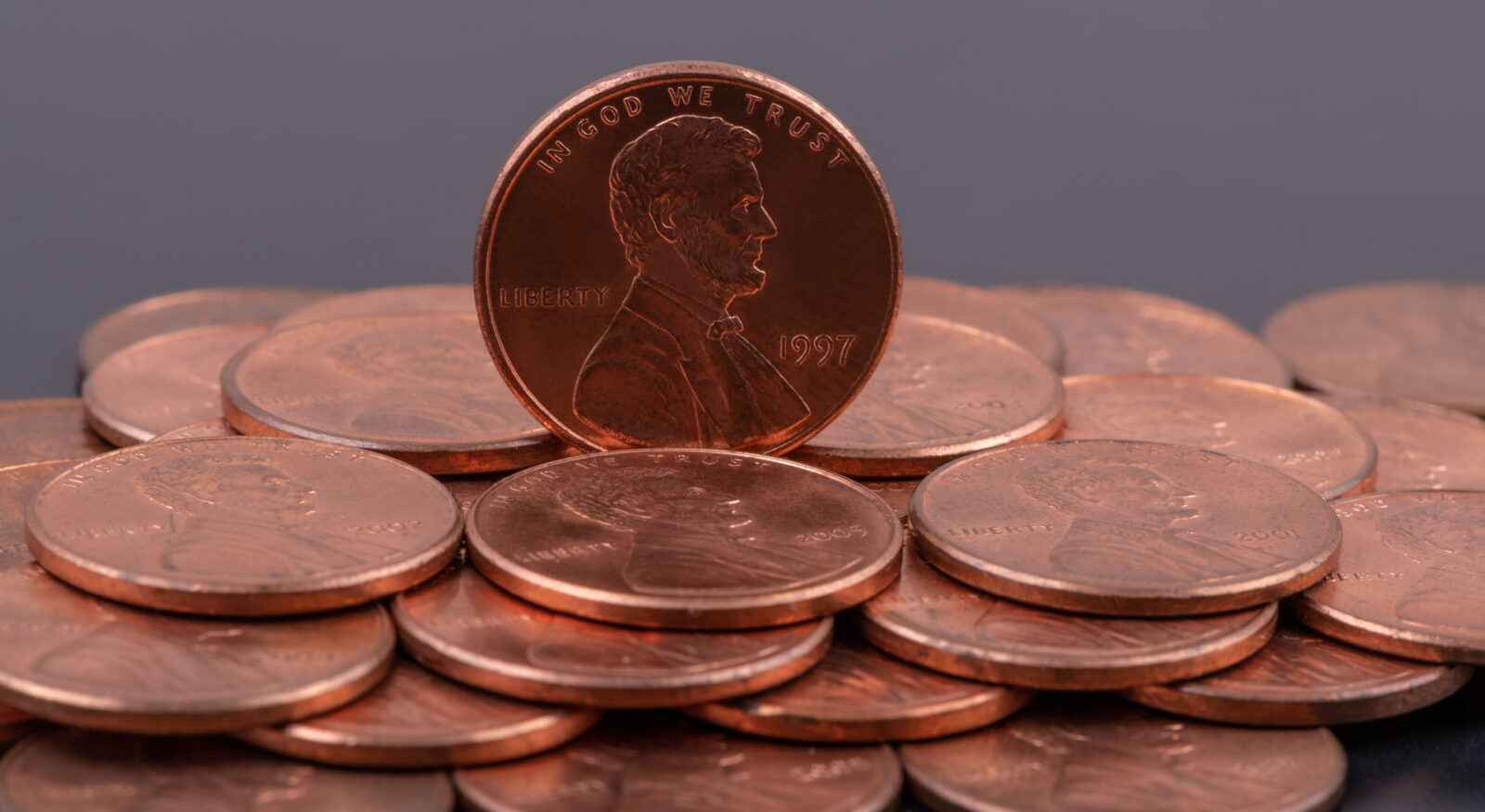 A group of U.S. one cent coins, with one 1997 penny standing upright among others lying flat on a dark background.