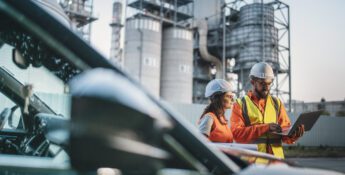 Two engineers in safety gear stand near industrial structures, reviewing information on a laptop beside a parked vehicle.