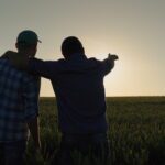 Two people stand in a field at sunset, one with an arm around the other and pointing towards the horizon, reflecting how farmers turning to peer groups seek support and share insights for a brighter future.