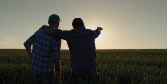 Two people stand in a field at sunset, one with an arm around the other and pointing towards the horizon, reflecting how farmers turning to peer groups seek support and share insights for a brighter future.