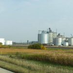 A large industrial facility with multiple silver storage tanks and silos is situated behind fields and shrubs under a partly cloudy sky.