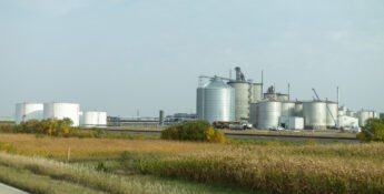 A large industrial facility with multiple silver storage tanks and silos is situated behind fields and shrubs under a partly cloudy sky.