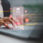 A person uses a laptop with a floating mobile banking login screen and multiple warning symbols, highlighting the importance of strengthening mobile banking defenses against cyber security threats.