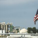An American flag flies in the foreground with an industrial plant featuring silos, towers, and storage tanks in the background—an apt scene as “One Big Beautiful Bill” introduces tax provisions that could boost biofuels under a cloudy sky.