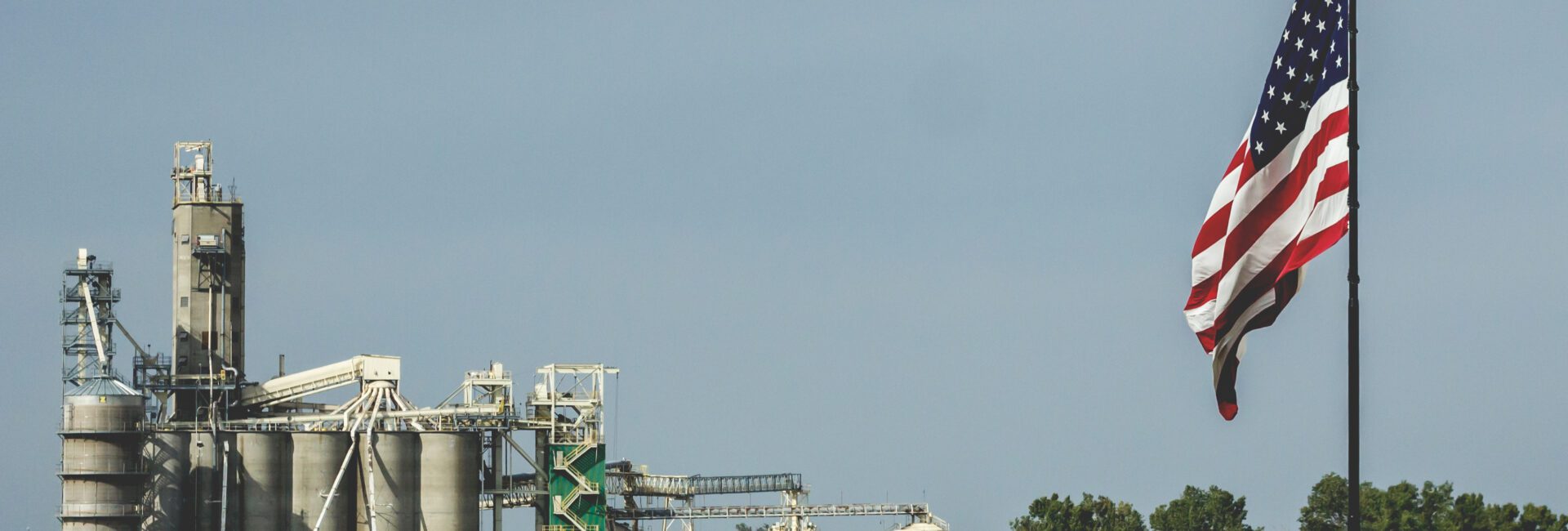 An American flag flies in the foreground with an industrial plant featuring silos, towers, and storage tanks in the background—an apt scene as “One Big Beautiful Bill” introduces tax provisions that could boost biofuels under a cloudy sky.