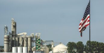 An American flag flies in the foreground with an industrial plant featuring silos, towers, and storage tanks in the background—an apt scene as “One Big Beautiful Bill” introduces tax provisions that could boost biofuels under a cloudy sky.