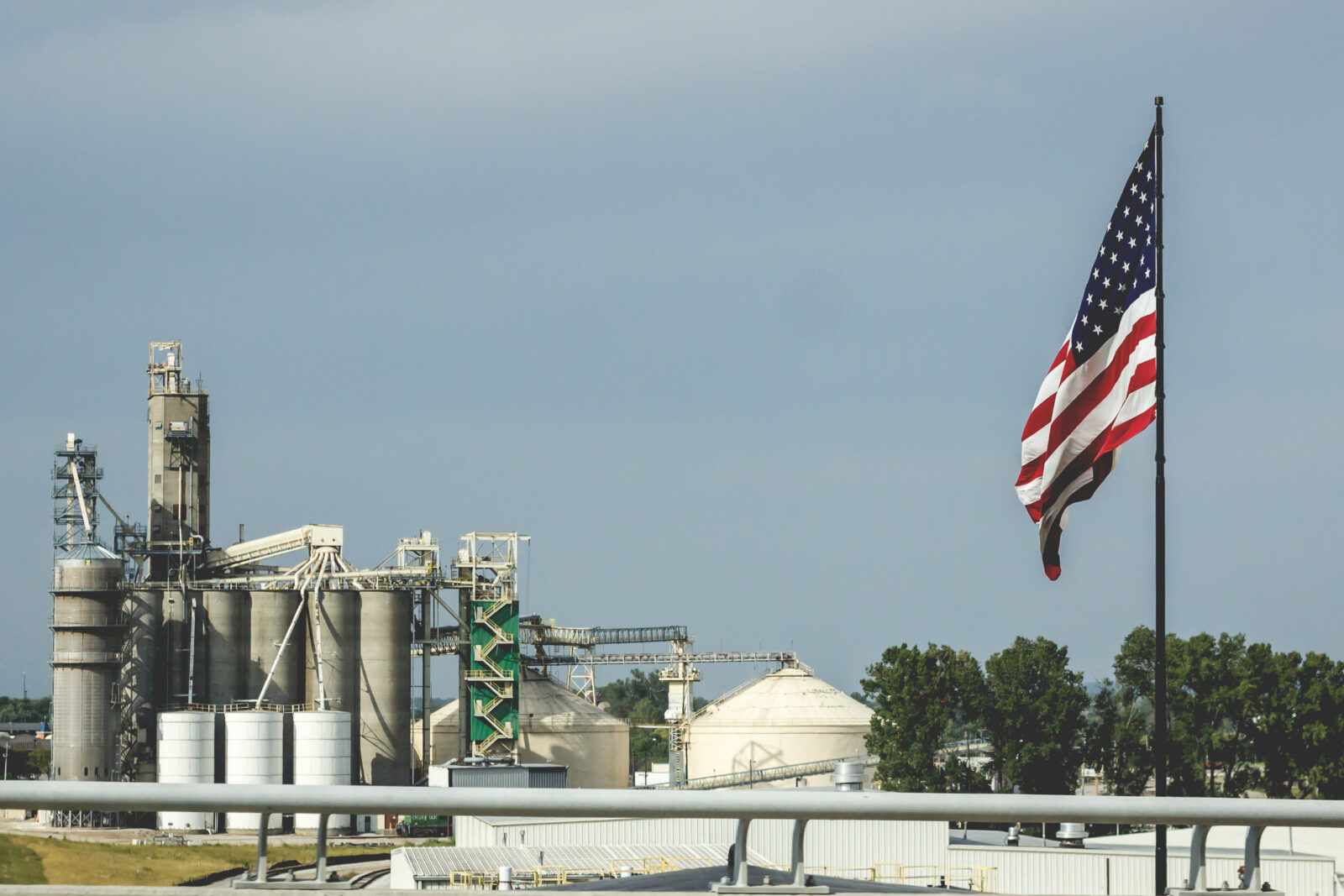 An American flag flies in the foreground with an industrial plant featuring silos, towers, and storage tanks in the background—an apt scene as “One Big Beautiful Bill” introduces tax provisions that could boost biofuels under a cloudy sky.