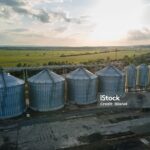 Aerial view of several large metal grain silos with surrounding infrastructure, set in a rural landscape under a partly cloudy sky at sunset, highlighting facilities preparing for 45Z clean fuel production credit.