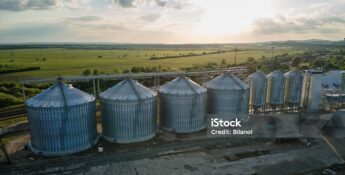 Aerial view of several large metal grain silos with surrounding infrastructure, set in a rural landscape under a partly cloudy sky at sunset, highlighting facilities preparing for 45Z clean fuel production credit.