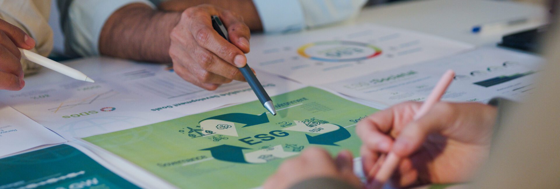 Three people discuss documents on a table, focusing on a green ESG (Environmental, Social, Governance) chart and exploring sustainability strategies with materiality assessments among other printed materials.