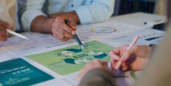 Three people discuss documents on a table, focusing on a green ESG (Environmental, Social, Governance) chart and exploring sustainability strategies with materiality assessments among other printed materials.