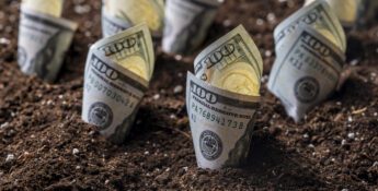 Several rolled-up US $100 bills are partially buried upright in dark soil, resembling small plants—an image that highlights key tips for surviving today’s ag economy.