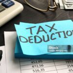A blue sticky note with "TAX DEDUCTION" written on it sits on financial documents, near a calculator, pen, glasses, and cash—highlighting the impact of New IRS Updates to Section 139L Loan Interest.