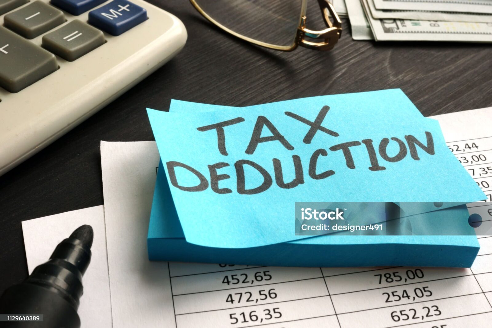 A blue sticky note with "TAX DEDUCTION" written on it sits on financial documents, near a calculator, pen, glasses, and cash—highlighting the impact of New IRS Updates to Section 139L Loan Interest.