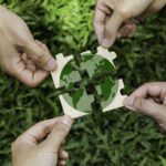 Four hands holding puzzle pieces that together form an image of the Earth, with green grass in the background—symbolizing global collaboration at the Sustainable Ag Summit.