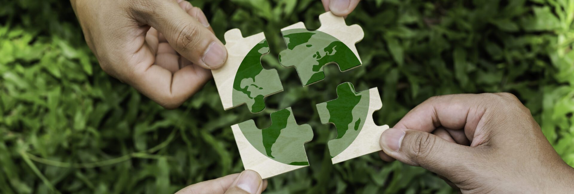 Four hands holding puzzle pieces that together form an image of the Earth, with green grass in the background—symbolizing global collaboration at the Sustainable Ag Summit.