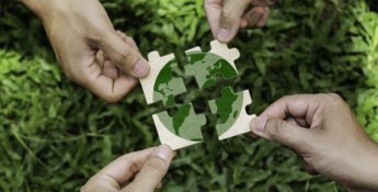 Four hands holding puzzle pieces that together form an image of the Earth, with green grass in the background—symbolizing global collaboration at the Sustainable Ag Summit.