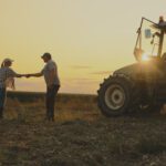 Two people shake hands in a field at sunset next to a tractor, with crops in the background—an inspiring moment that reflects the collaborative spirit of Sustainability Peer Groups in agriculture.
