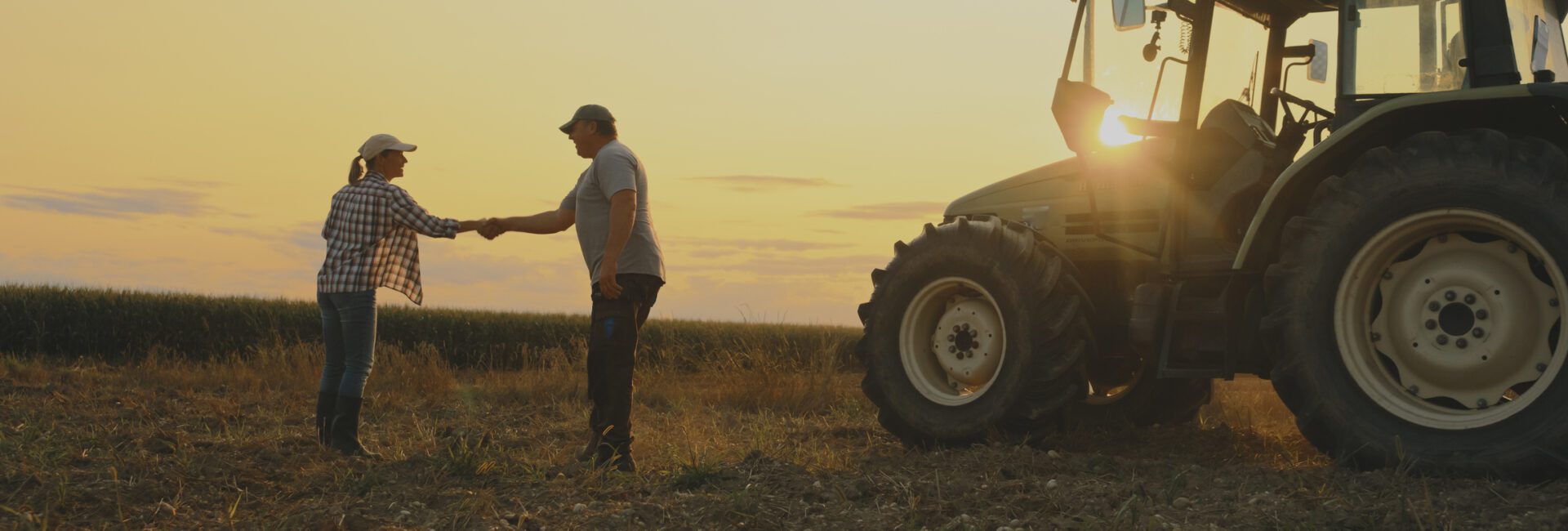 Two people shake hands in a field at sunset next to a tractor, with crops in the background—an inspiring moment that reflects the collaborative spirit of Sustainability Peer Groups in agriculture.