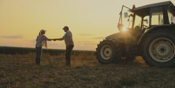 Two people shake hands in a field at sunset next to a tractor, with crops in the background—an inspiring moment that reflects the collaborative spirit of Sustainability Peer Groups in agriculture.