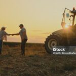 Two people shake hands in a field at sunset, standing next to a tractor, embodying the spirit of Sustainability Peer Groups. Crops and sky form a scenic background, highlighting collaboration for a greener future.