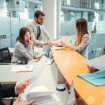 Five people in business attire work at a modern office desk, reviewing documents, using laptops, and having discussions in a bright, glass-walled space.