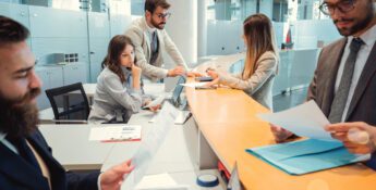 Five people in business attire work at a modern office desk, reviewing documents, using laptops, and having discussions in a bright, glass-walled space.