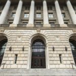 The image shows the front entrance of a large neoclassical building with tall stone columns, arched doorways, and wide steps leading up to the doors.