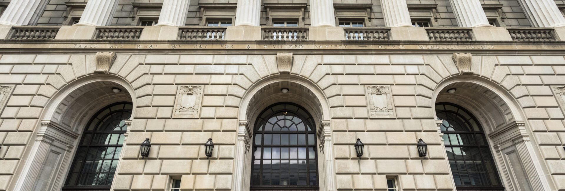 The image shows the front entrance of a large neoclassical building with tall stone columns, arched doorways, and wide steps leading up to the doors.
