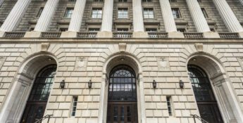 The image shows the front entrance of a large neoclassical building with tall stone columns, arched doorways, and wide steps leading up to the doors.