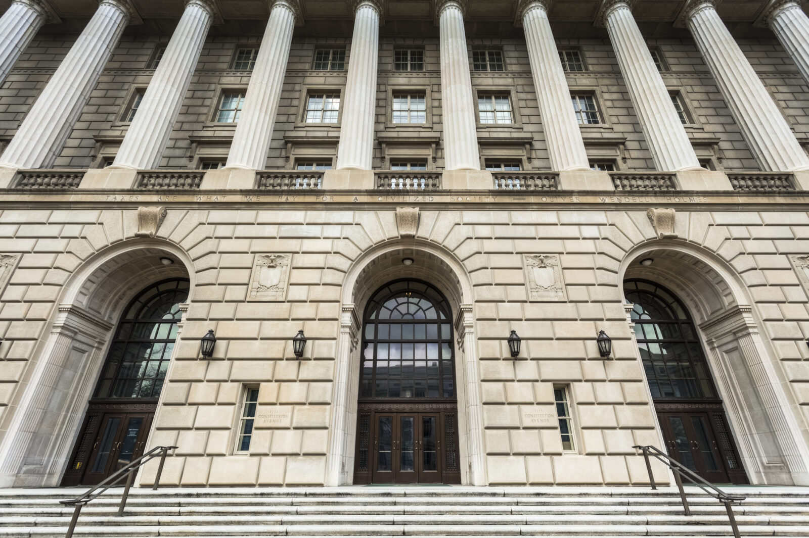 The image shows the front entrance of a large neoclassical building with tall stone columns, arched doorways, and wide steps leading up to the doors.