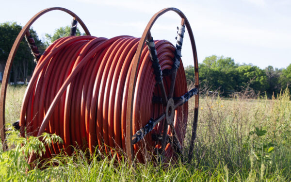 A large spool of red hose or tubing is situated in a grassy field with trees in the background.