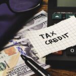 A calculator, US $100 bills, a pen, a notebook, and a piece of paper labeled "TAX CREDIT" highlight the importance of the Work Opportunity Tax Credit: Why Employers Should Keep Filing on your desk.