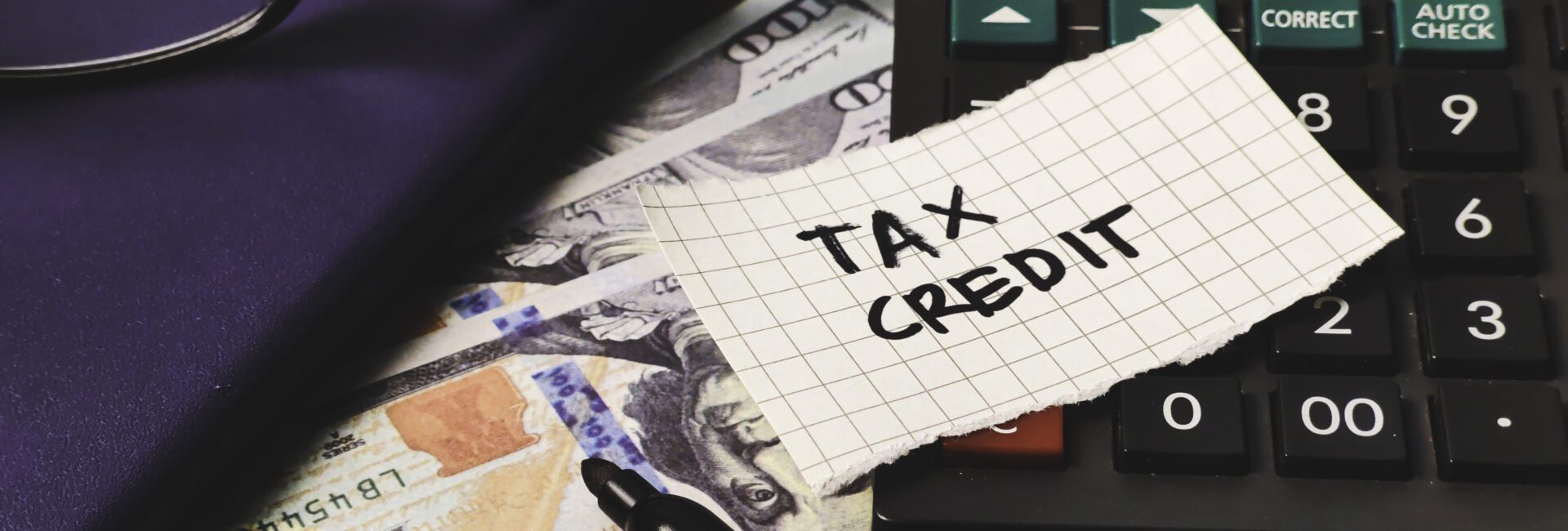 A calculator, US $100 bills, a pen, a notebook, and a piece of paper labeled "TAX CREDIT" highlight the importance of the Work Opportunity Tax Credit: Why Employers Should Keep Filing on your desk.