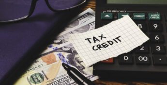 A calculator, US $100 bills, a pen, a notebook, and a piece of paper labeled "TAX CREDIT" highlight the importance of the Work Opportunity Tax Credit: Why Employers Should Keep Filing on your desk.