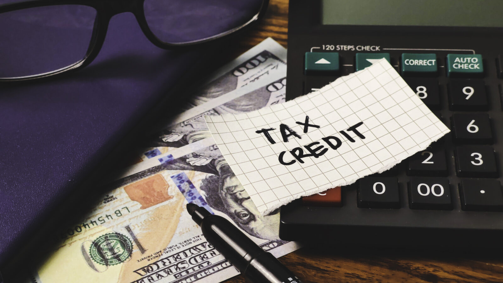 A calculator, US $100 bills, a pen, a notebook, and a piece of paper labeled "TAX CREDIT" highlight the importance of the Work Opportunity Tax Credit: Why Employers Should Keep Filing on your desk.