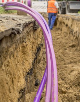 Two large purple pipes are laid in a sand trench at a construction site, with a worker in a safety vest and helmet standing nearby.