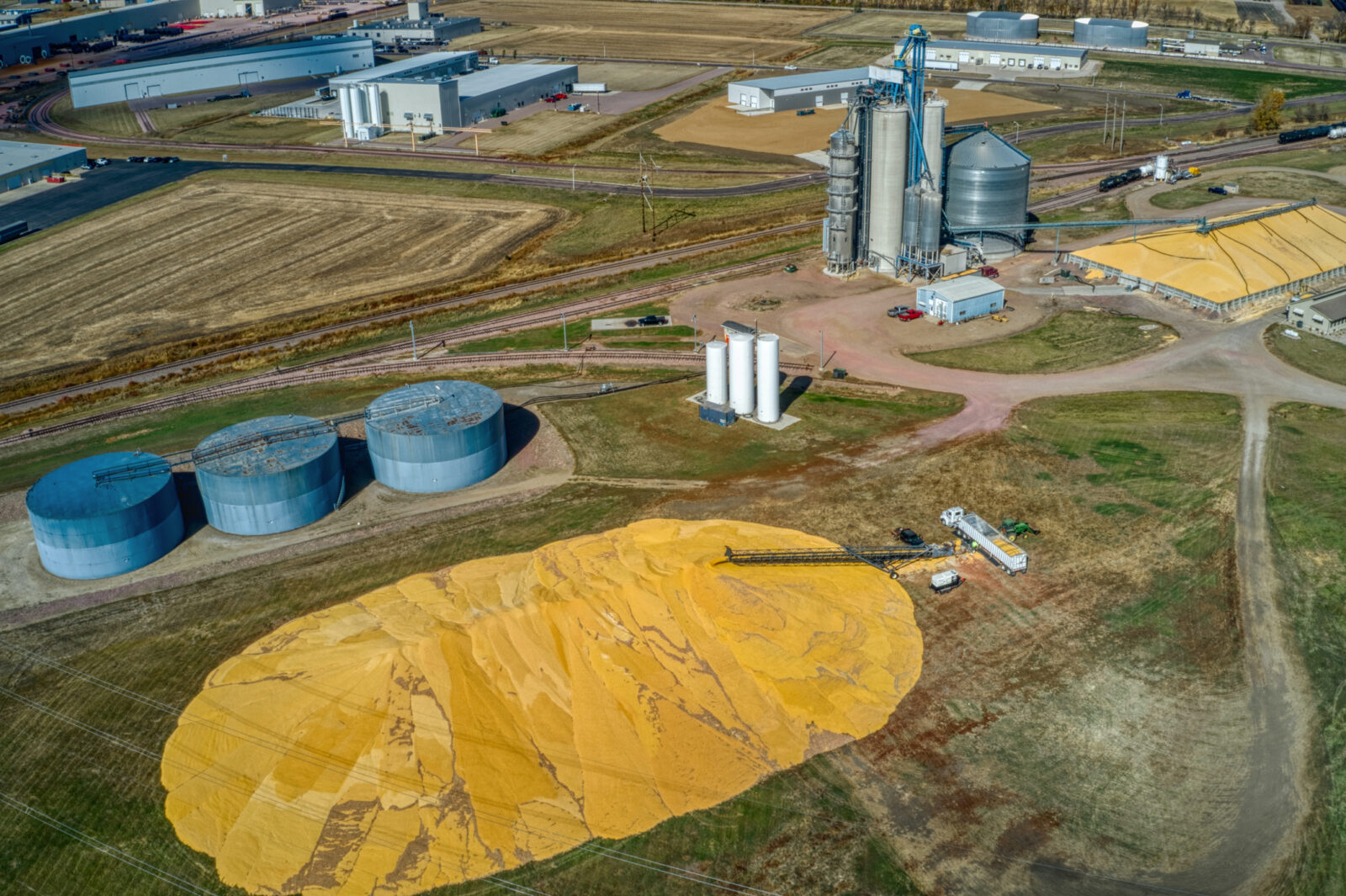 Aerial view of an agricultural facility with large grain silos, storage tanks, and piles of harvested grain spread on the ground, illustrating preparations for biofuels volatility in today's evolving energy landscape.