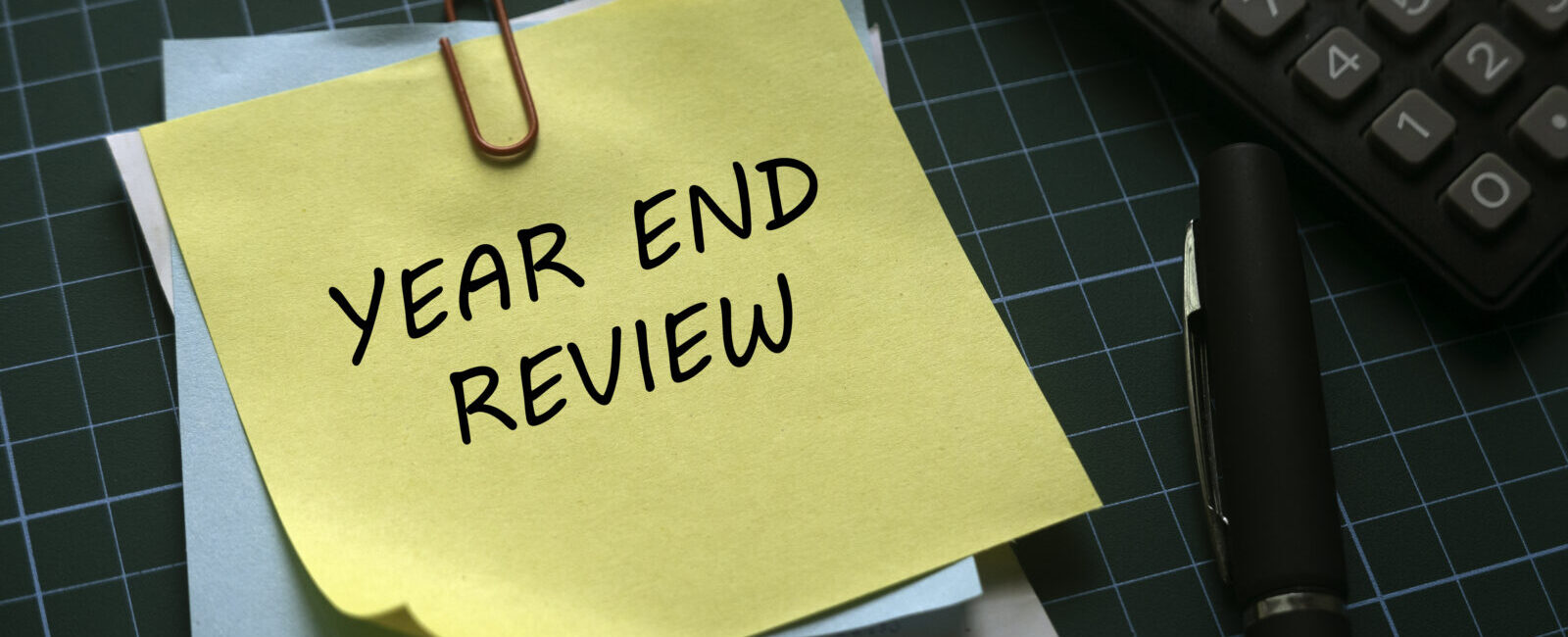 A yellow sticky note with "YEAR END REVIEW" written on it is clipped to paperwork beside a pen and calculator on a green cutting mat—perfect for turning your nonprofit year-end into a strategic advantage.