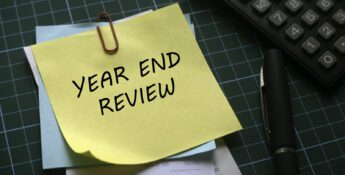 A yellow sticky note with "YEAR END REVIEW" written on it is clipped to paperwork beside a pen and calculator on a green cutting mat—perfect for turning your nonprofit year-end into a strategic advantage.