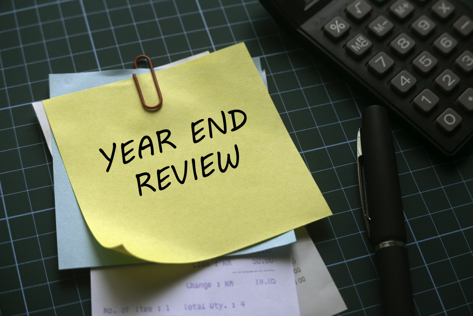 A yellow sticky note with "YEAR END REVIEW" written on it is clipped to paperwork beside a pen and calculator on a green cutting mat—perfect for turning your nonprofit year-end into a strategic advantage.