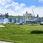 An industrial facility with large metal storage tanks and various structures sits on a grassy area under a partly cloudy sky.