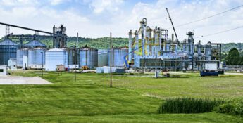 An industrial facility with large metal storage tanks and various structures sits on a grassy area under a partly cloudy sky.