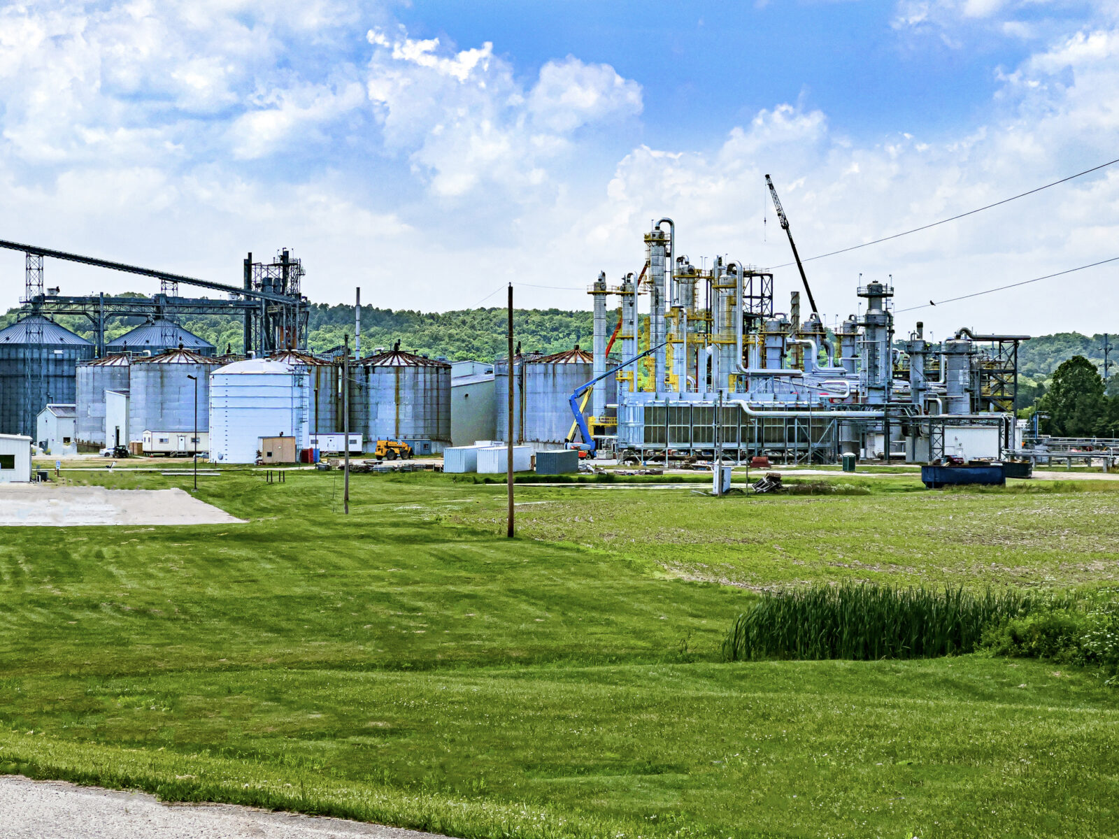 An industrial facility with large metal storage tanks and various structures sits on a grassy area under a partly cloudy sky.