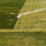 Aerial view of a tractor harvesting a large green field, leaving visible lines and a trail of dust behind.